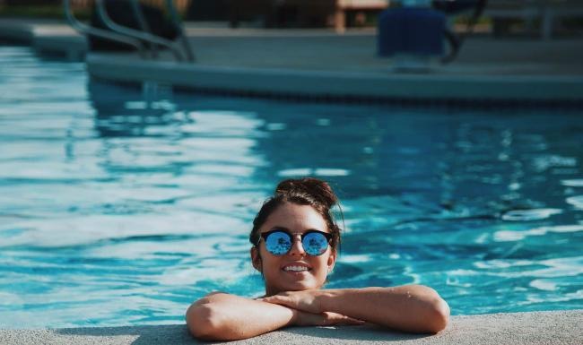 woman with sunglasses, in a pool, rests her arms on the ledge and smiles